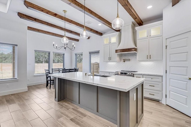 kitchen featuring pendant lighting, white cabinetry, premium range hood, sink, and a kitchen island with sink