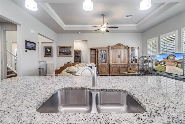 kitchen with light stone counters, a raised ceiling, ceiling fan, sink, and hanging light fixtures