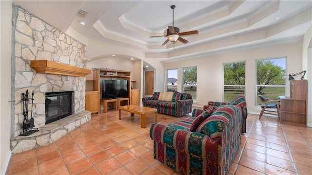 tiled living room featuring ceiling fan, a stone fireplace, a raised ceiling, and ornamental molding