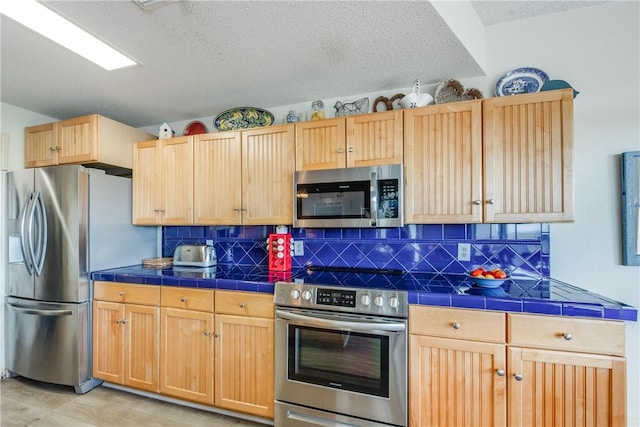 kitchen with appliances with stainless steel finishes, tile counters, a textured ceiling, light brown cabinets, and backsplash