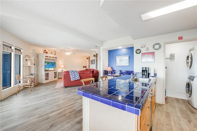 kitchen featuring a sink, stacked washing machine and dryer, tile counters, light wood-style floors, and a textured ceiling