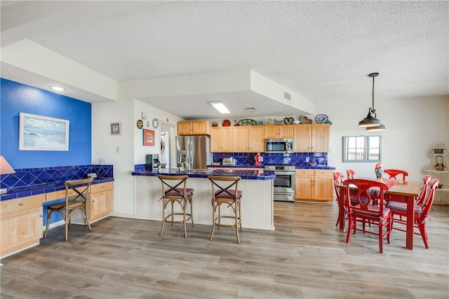 kitchen with stainless steel appliances, light wood finished floors, decorative backsplash, light brown cabinetry, and a textured ceiling