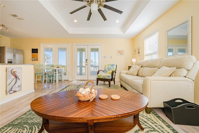 living room with a raised ceiling, ceiling fan, french doors, and light wood-type flooring