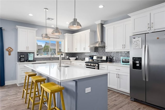 kitchen with wall chimney range hood, hanging light fixtures, stainless steel appliances, white cabinets, and a center island with sink