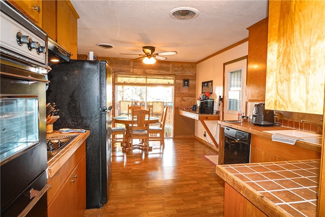 kitchen featuring tile countertops, black appliances, ornamental molding, ceiling fan, and light wood-type flooring