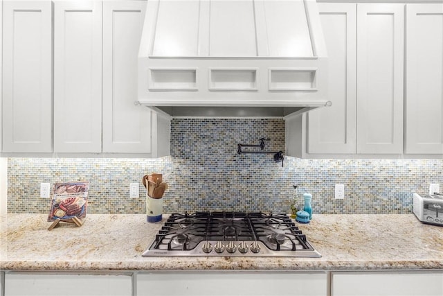 kitchen featuring light stone counters, tasteful backsplash, stainless steel gas stovetop, white cabinets, and premium range hood