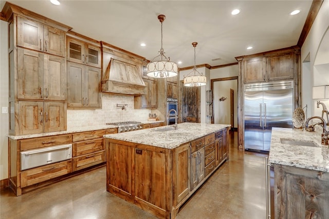 kitchen featuring appliances with stainless steel finishes, light stone counters, custom range hood, a kitchen island with sink, and decorative light fixtures