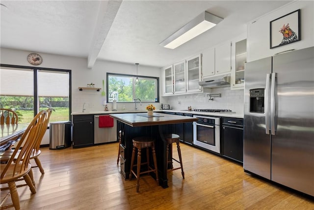 kitchen featuring under cabinet range hood, beamed ceiling, light wood-style flooring, tasteful backsplash, and appliances with stainless steel finishes