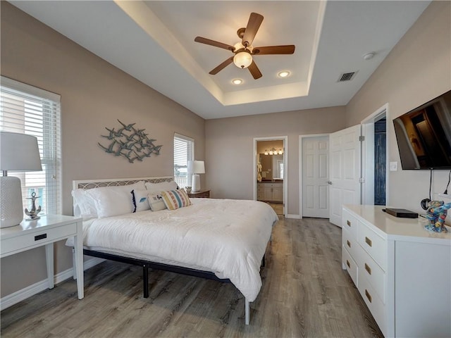 bedroom featuring ceiling fan, ensuite bath, light wood-type flooring, and a raised ceiling