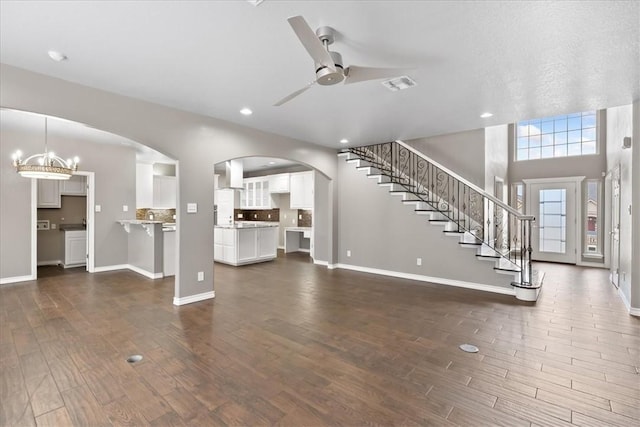 unfurnished living room featuring ceiling fan with notable chandelier, visible vents, baseboards, stairs, and dark wood finished floors