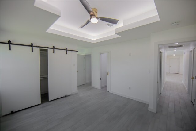 unfurnished bedroom featuring a barn door, light hardwood / wood-style floors, ceiling fan, and a raised ceiling