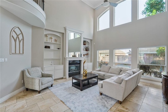 living area with a towering ceiling, built in shelves, baseboards, ceiling fan, and a glass covered fireplace