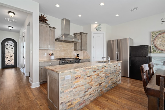 kitchen with wall chimney range hood, appliances with stainless steel finishes, light stone countertops, dark wood-type flooring, and a kitchen island with sink