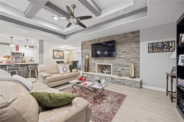 living room featuring ceiling fan, a tray ceiling, a stone fireplace, and light hardwood / wood-style flooring