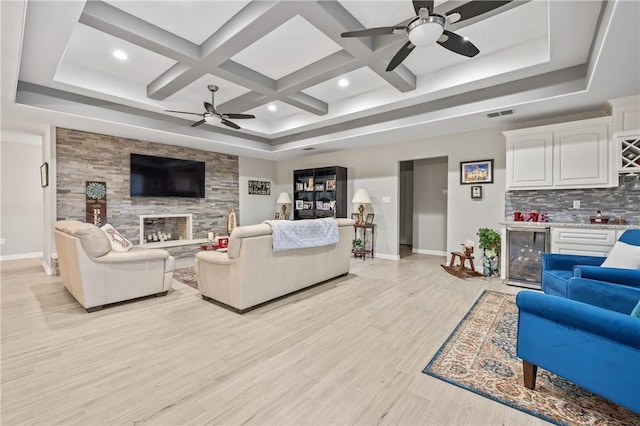 living room with ceiling fan, coffered ceiling, a stone fireplace, beverage cooler, and light wood-type flooring