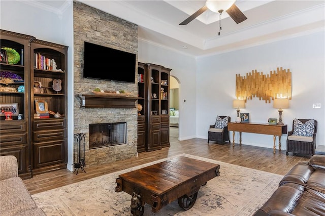 living area featuring arched walkways, ceiling fan, a stone fireplace, wood finished floors, and ornamental molding