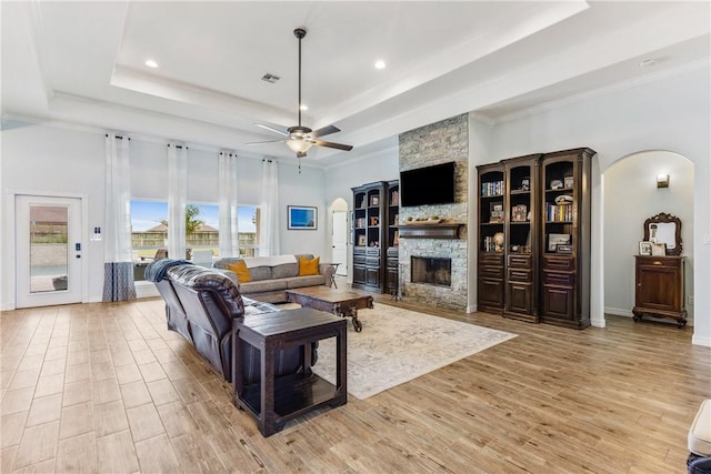 living room with light wood finished floors, arched walkways, a raised ceiling, and a stone fireplace