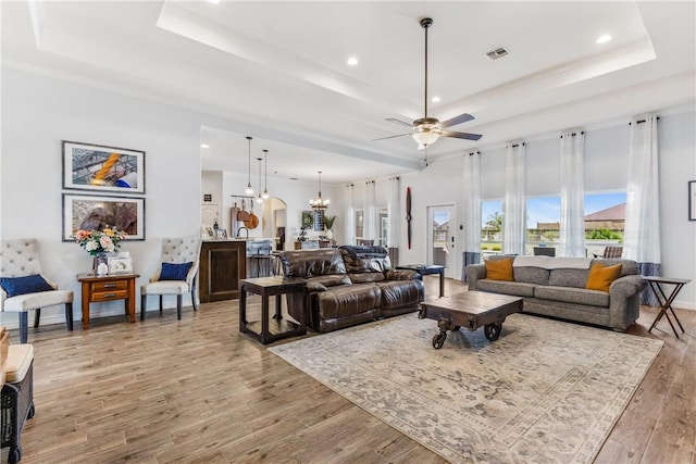 living room featuring a tray ceiling, visible vents, light wood finished floors, and ceiling fan with notable chandelier