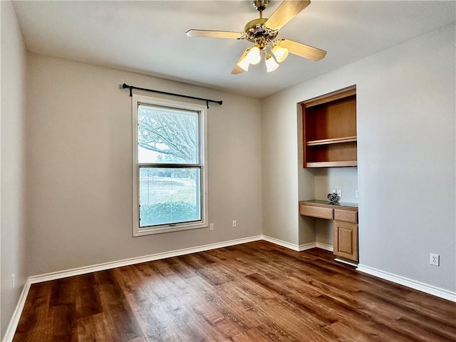 spare room featuring dark wood-type flooring, built in study area, ceiling fan, and baseboards