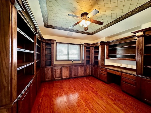 unfurnished office with dark wood-type flooring, a tray ceiling, ceiling fan, and built in desk