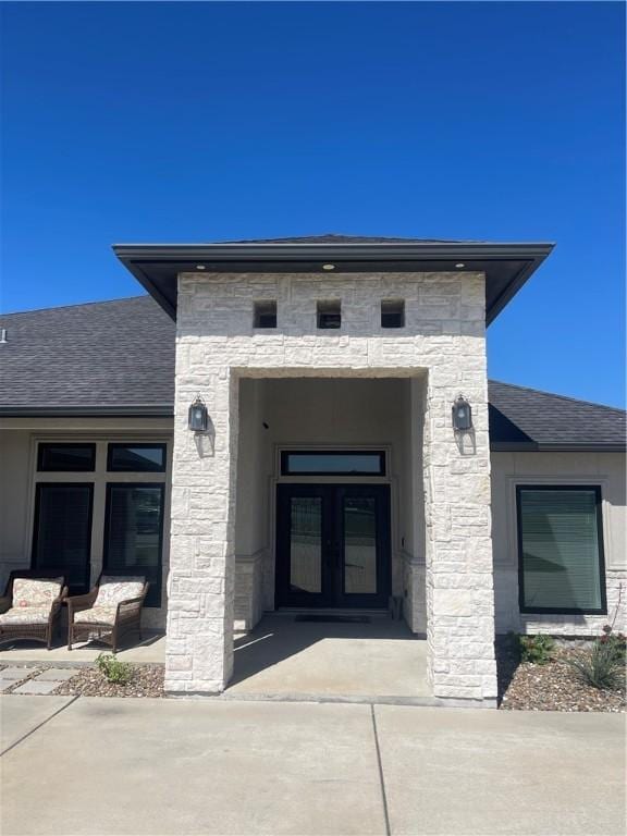 view of exterior entry with stone siding, french doors, and roof with shingles