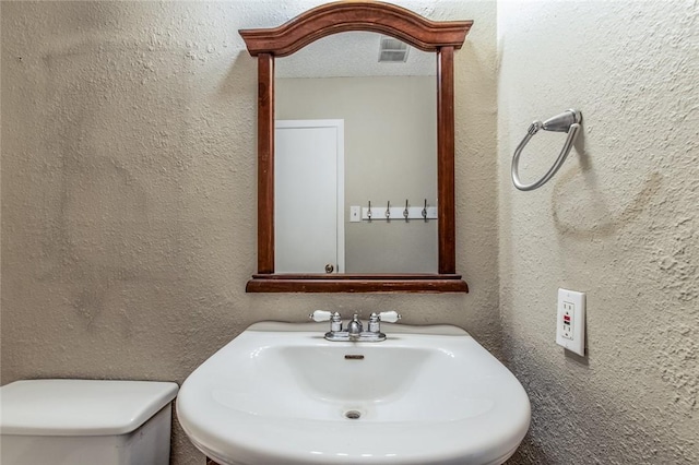 bathroom featuring toilet, a textured wall, visible vents, and a sink