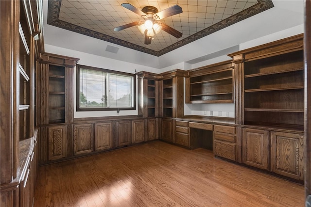 unfurnished office with a tray ceiling, built in desk, ceiling fan, and light hardwood / wood-style flooring