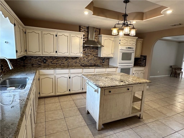 kitchen featuring sink, light stone counters, hanging light fixtures, a kitchen island, and wall chimney range hood