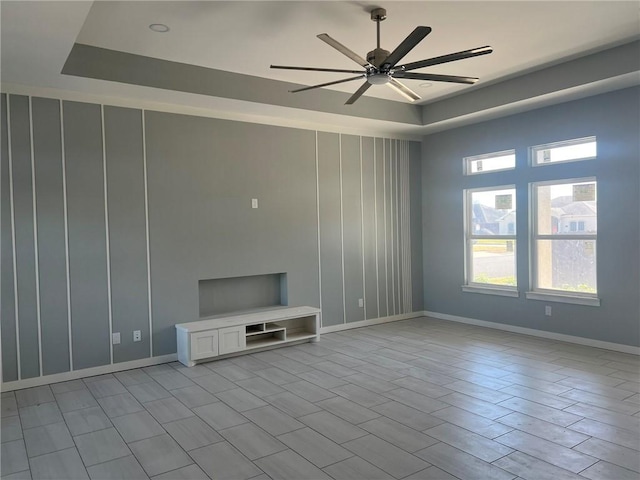 unfurnished living room with ceiling fan, a raised ceiling, a wealth of natural light, and baseboards