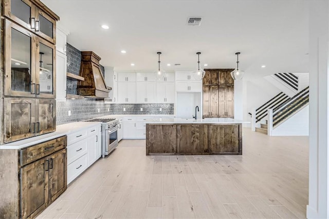 kitchen with white cabinets, backsplash, hanging light fixtures, stainless steel range, and a center island with sink