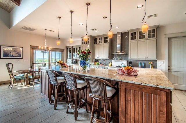 kitchen with backsplash, wall chimney range hood, decorative light fixtures, a large island with sink, and light stone countertops