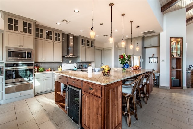 kitchen featuring a center island with sink, stainless steel appliances, wine cooler, wall chimney range hood, and decorative light fixtures