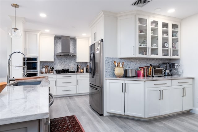 kitchen with light stone counters, stainless steel fridge, wall chimney range hood, hanging light fixtures, and white cabinets