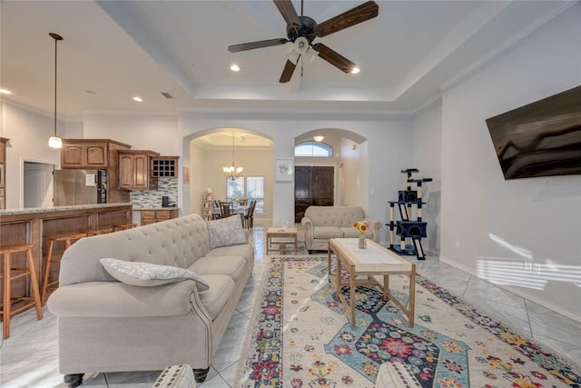 tiled living room featuring a raised ceiling, crown molding, and ceiling fan with notable chandelier