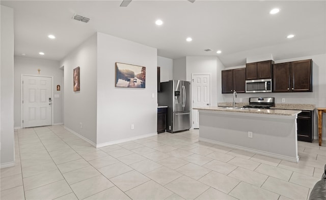 kitchen with an island with sink, light stone counters, appliances with stainless steel finishes, and dark brown cabinetry