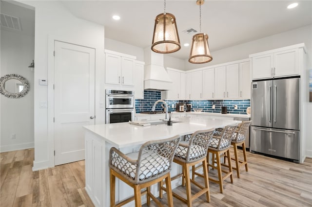 kitchen with stainless steel appliances, premium range hood, white cabinetry, light countertops, and a center island with sink