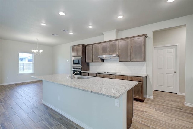 kitchen featuring a kitchen island with sink, appliances with stainless steel finishes, a notable chandelier, light stone counters, and tasteful backsplash