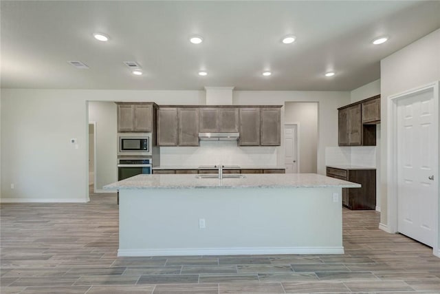 kitchen with a kitchen island with sink, appliances with stainless steel finishes, light stone counters, sink, and backsplash