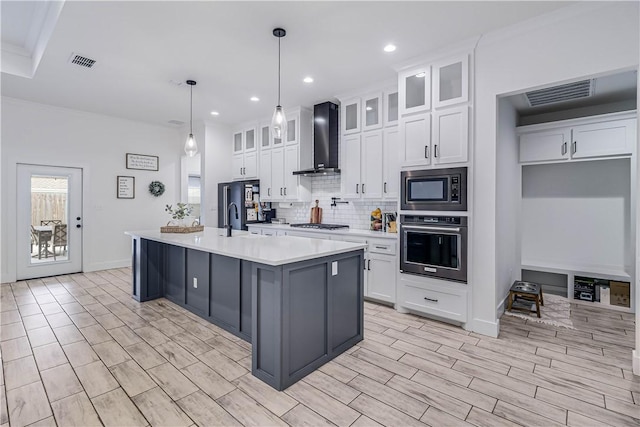 kitchen with decorative light fixtures, wall chimney range hood, white cabinetry, an island with sink, and stainless steel appliances