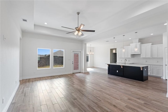 kitchen with ceiling fan with notable chandelier, a tray ceiling, pendant lighting, white cabinetry, and an island with sink