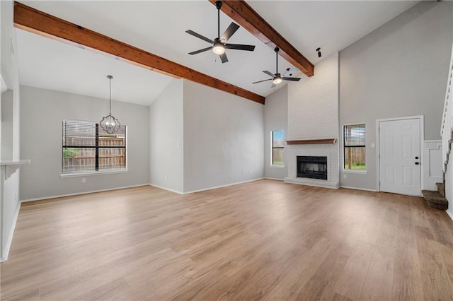 unfurnished living room with ceiling fan with notable chandelier, high vaulted ceiling, a fireplace, beamed ceiling, and light hardwood / wood-style flooring