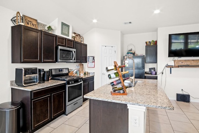 kitchen featuring an island with sink, appliances with stainless steel finishes, dark brown cabinets, light tile patterned flooring, and light stone counters