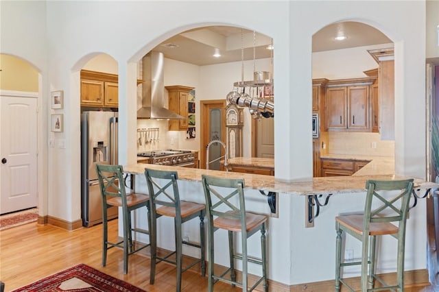 kitchen with light wood-style floors, appliances with stainless steel finishes, light stone countertops, wall chimney exhaust hood, and a kitchen bar