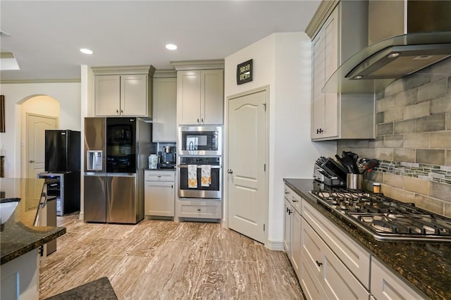 kitchen featuring appliances with stainless steel finishes, backsplash, wall chimney exhaust hood, and dark stone countertops