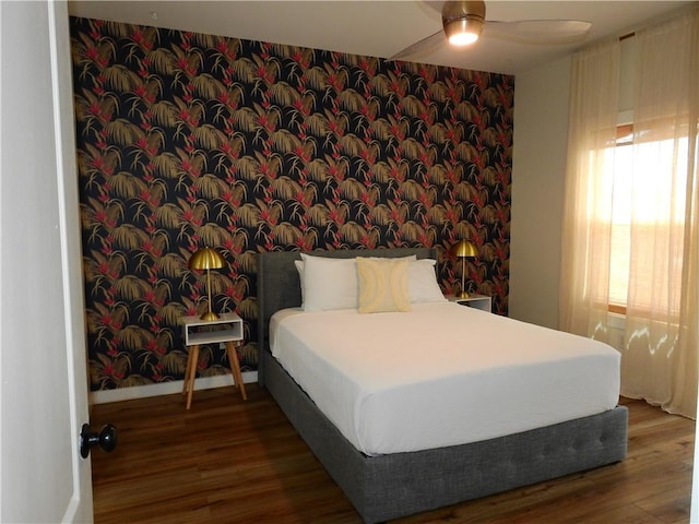 bedroom featuring ceiling fan, an accent wall, wood finished floors, baseboards, and wallpapered walls