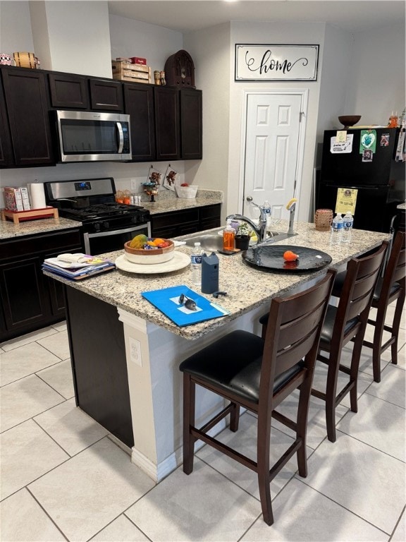 kitchen with stainless steel appliances, a center island with sink, light stone counters, light tile patterned floors, and a breakfast bar