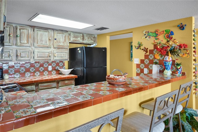 kitchen with tile counters, a textured ceiling, black fridge, and tasteful backsplash