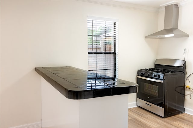 kitchen with tile counters, stainless steel gas stove, wall chimney exhaust hood, light hardwood / wood-style flooring, and ornamental molding