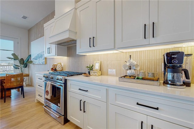 kitchen with tasteful backsplash, white cabinetry, custom exhaust hood, light hardwood / wood-style floors, and stainless steel gas range oven