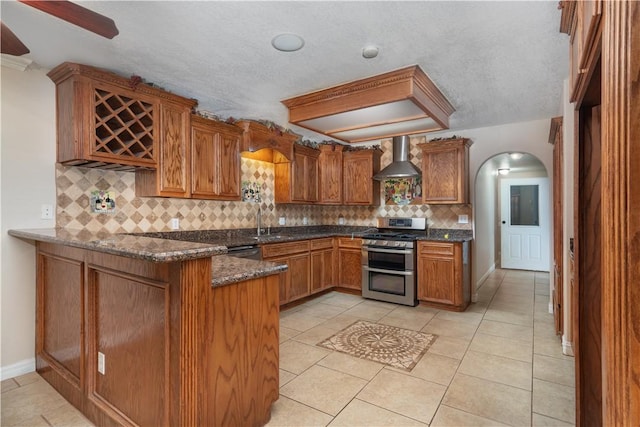 kitchen with kitchen peninsula, decorative backsplash, wall chimney exhaust hood, stainless steel appliances, and dark stone countertops